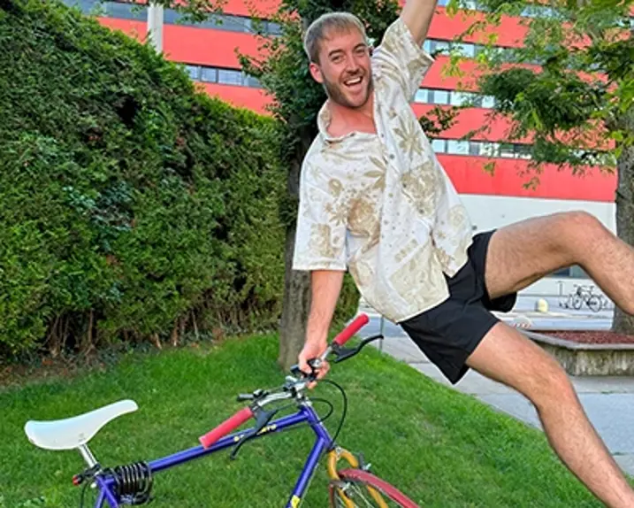 Ein Influencer mit dem Namen Jonny hängt an einem Baum knapp über dem Boden. Er hält mit der rechten Hand ein buntes Fahrrad. Im Hintergrund sieht man ein Haus und Wiese. Jonny trägt eine Kurzarmhemd, eine kurze Hose.