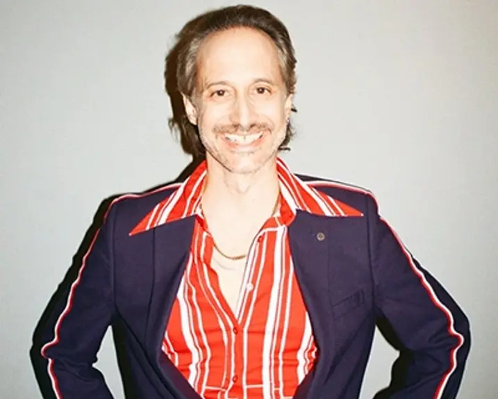 Der Schauspieler Michael Ostrowski lächelt in die Kamera. Er trägt ein rot-weiß-gestreiftes Hemd darüber eine Art Blouson in dunkelblau. Dieser hat ebenfalls einen roten und einen weißen Streifen im Bereich der Oberarme.